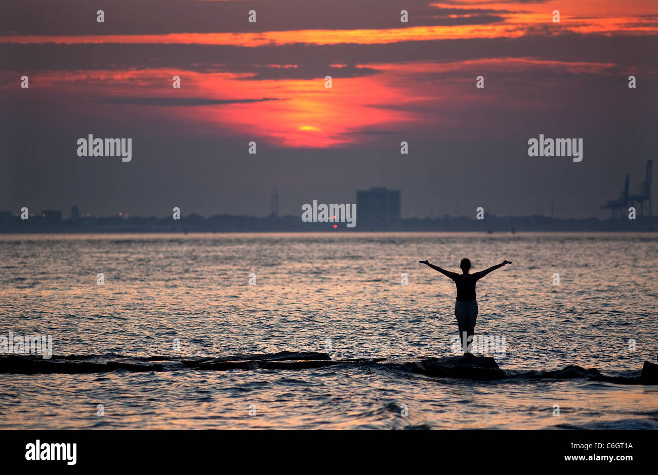 Une femme danse sur rock jetées au large de l'Île Sullivan en Caroline du Sud avec Charleston dans l'arrière-plan pendant le coucher du soleil. Banque D'Images