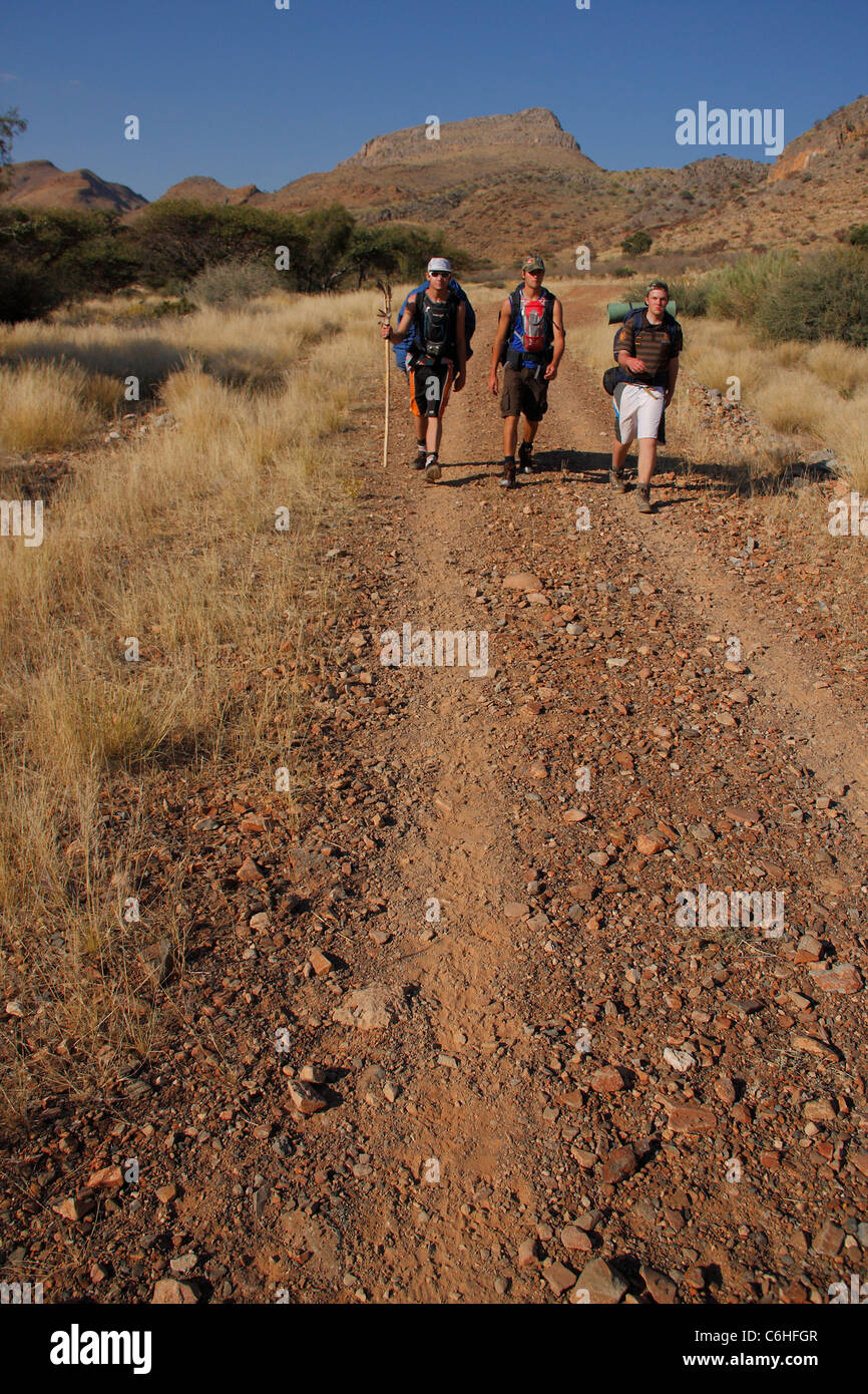 Trois randonneurs marchant le long d'une route de gravier dans paysage de désert Banque D'Images