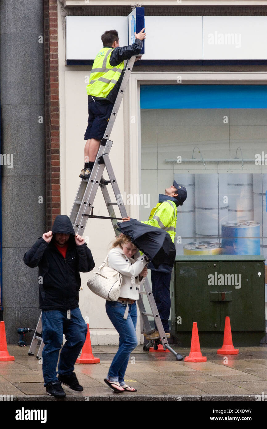 Jeune couple lutte dans le vent et la pluie comme ils passent deux ouvriers avec une échelle Banque D'Images