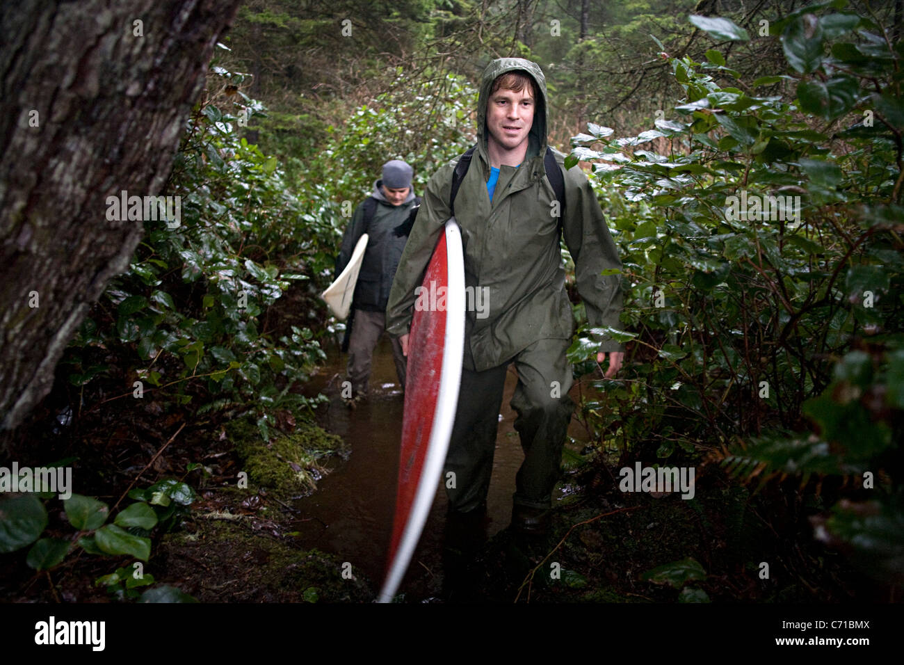 Homme portant des vêtements de pluie est titulaire d'une planche de surf sous le bras dans la forêt à pied Banque D'Images