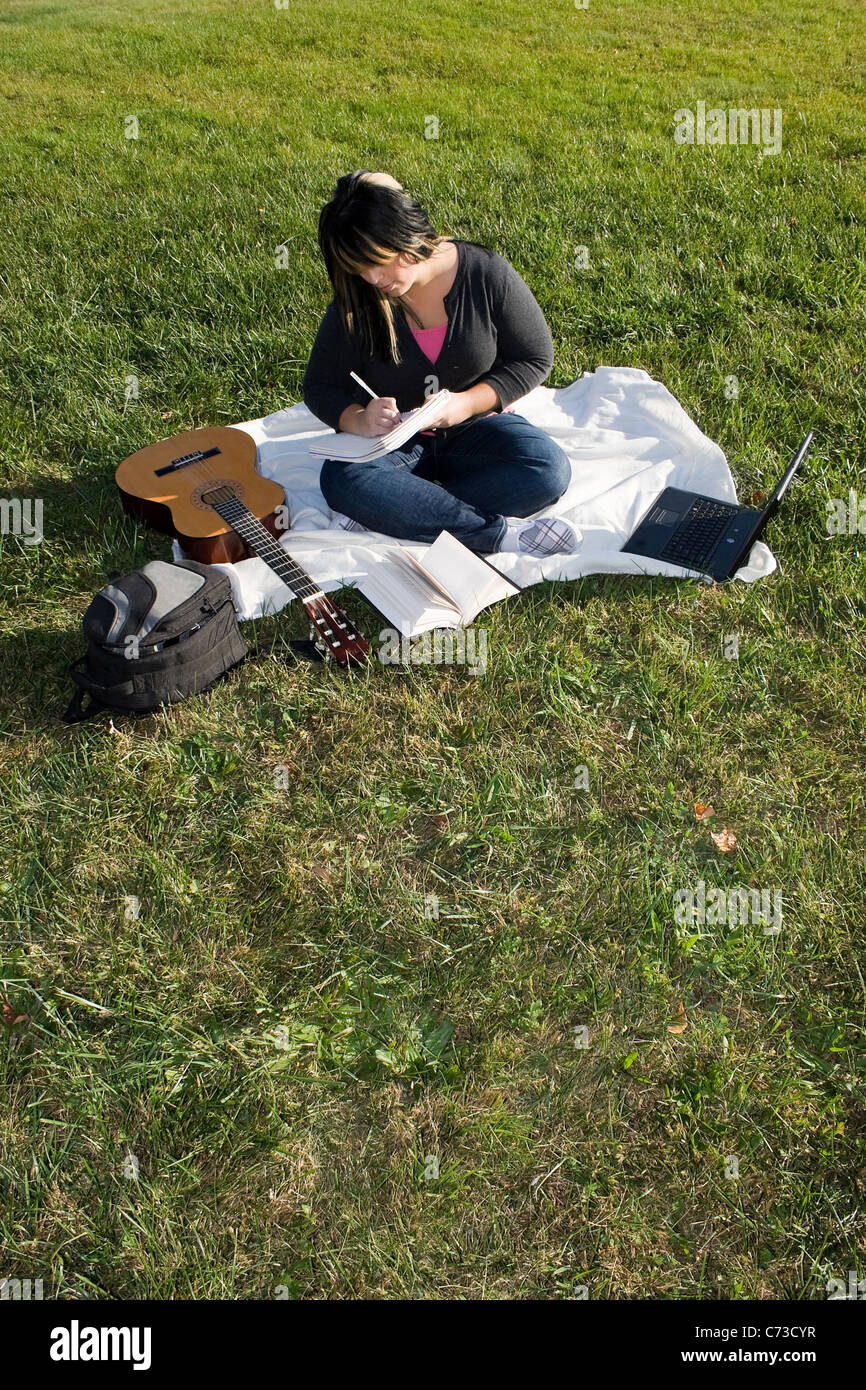 Un jeune musicien écrit dans son ordinateur portable tout en étant assis dans l'herbe par une belle journée. Banque D'Images