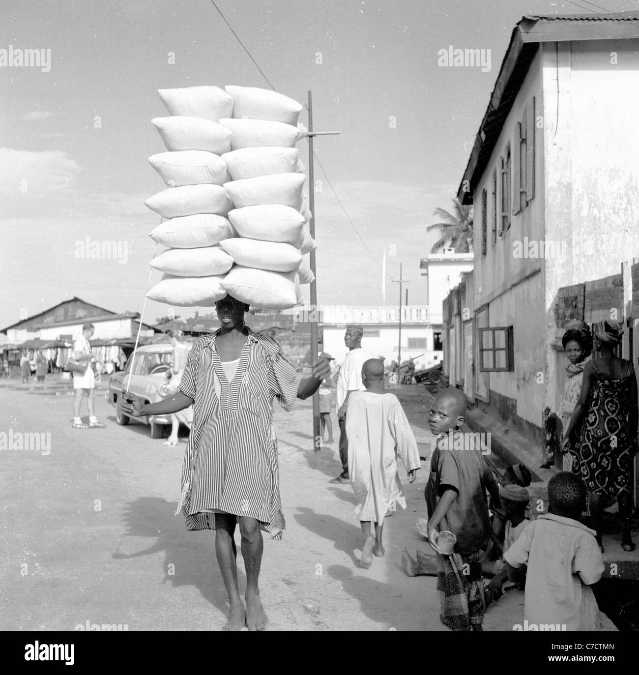 Le Nigeria. Années 1950. Dame locale walking down street abilement en balance une grande charge de nourriture sacs ou des paquets sur la tête, Banque D'Images