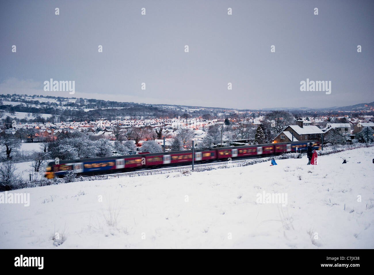 Soirée hivernale enneigée, train de passagers qui passe devant le village rural et le champ couvert de neige blanche - ligne Wharfedale, West Yorkshire, Angleterre, Royaume-Uni. Banque D'Images