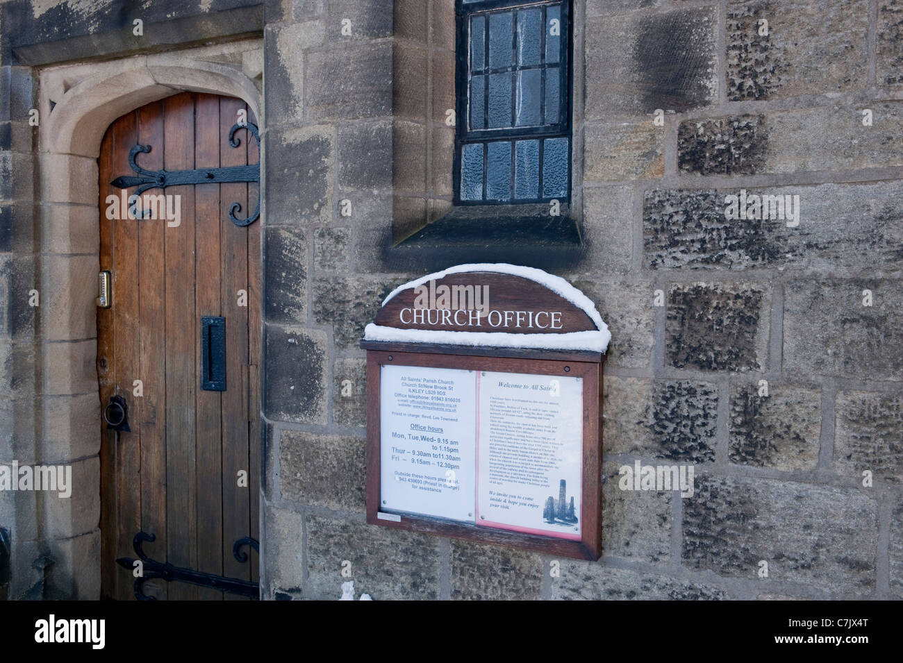 Extérieur du bureau de l'église, fermeture de la porte d'entrée en bois (charnières à sangle) mur en pierre et neige sur le panneau de remarque - Église paroissiale de tous les Saints, Ilkley, Angleterre, Royaume-Uni. Banque D'Images