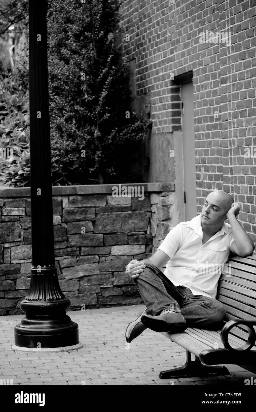 Un homme d'une vingtaine d'années assis nonchalamment sur un banc dans une zone urbaine. Banque D'Images