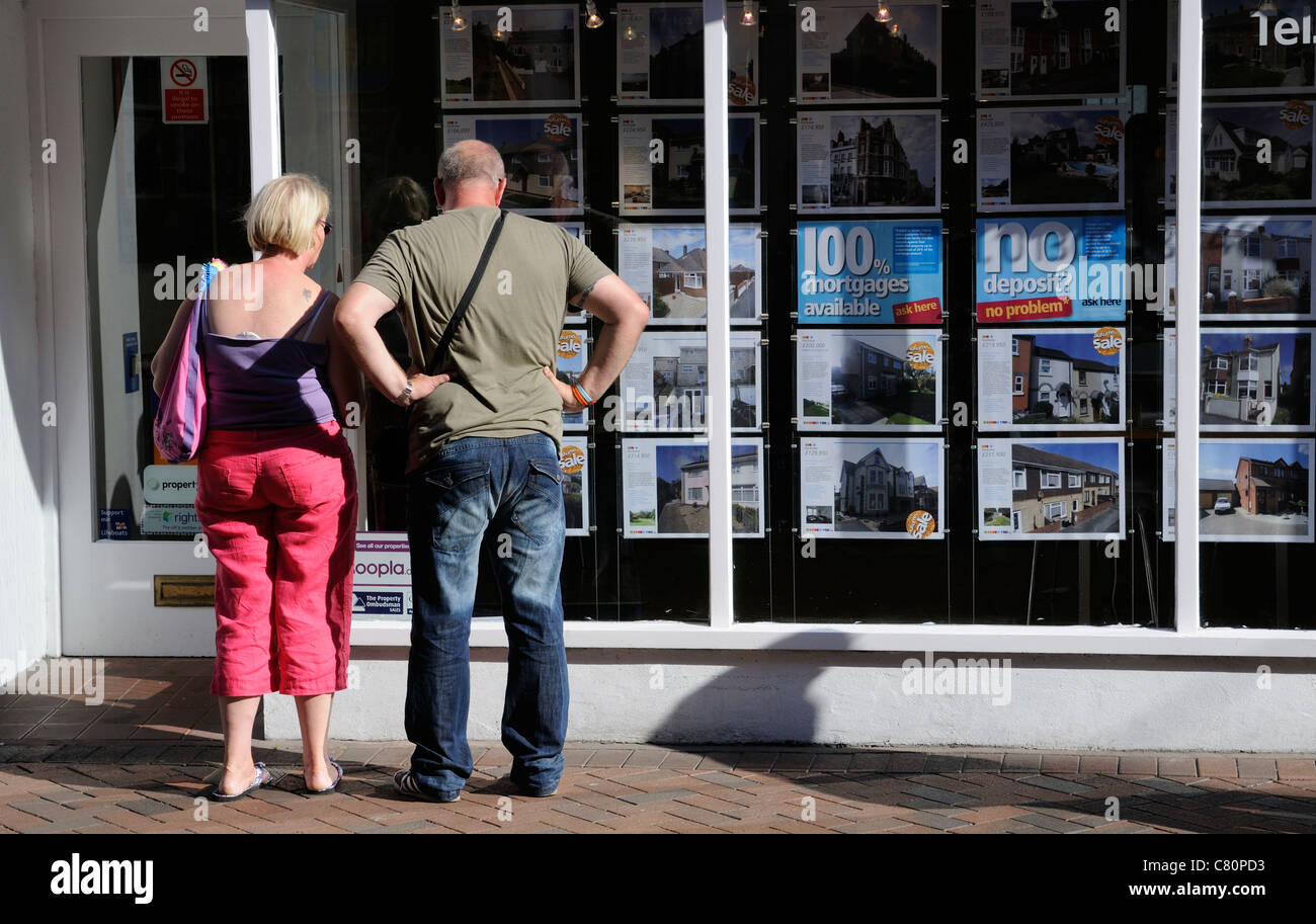 Couple recherchant la propriété à vendre à repasser dans une fenêtre d'agents immobiliers Banque D'Images