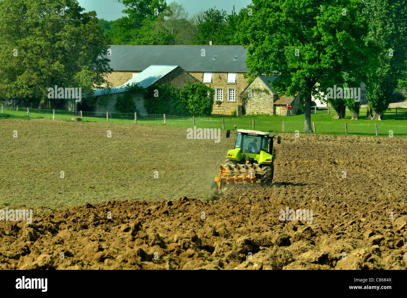 Un tracteur laboure un champ, une ferme en arrière-plan (Mayenne, Loire, France). Banque D'Images