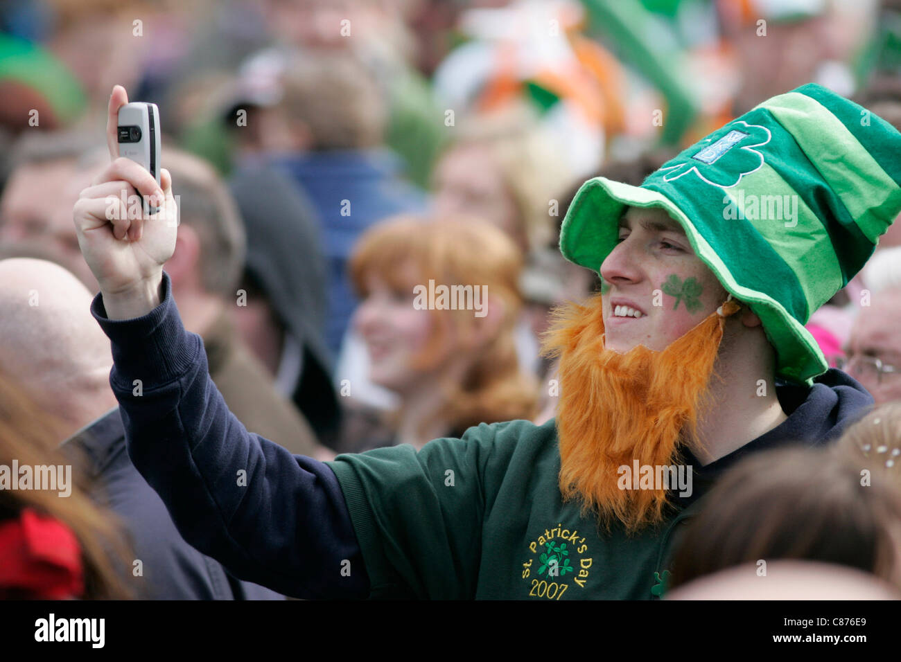 Jeune homme avec barbe orange faux prend un téléphone mobile photo dans la foule sur St Patricks Day Banque D'Images