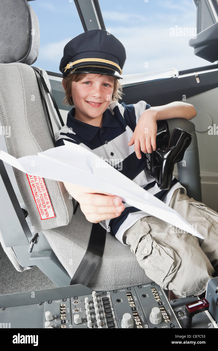 Germany, Bavaria, Munich, Boy wearing captain's hat and holding paper plane in airplane cockpit Banque D'Images