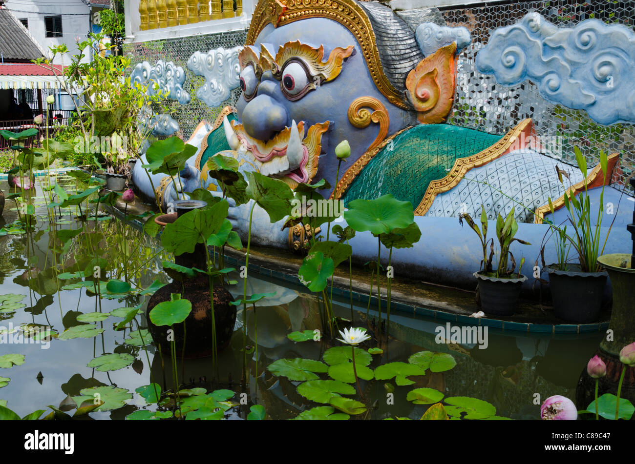 Statue de divinité bouddhiste coloré Kala avec yeux exorbités et un étang à l'avant avec la floraison de nénuphars au temple thaïlandais Banque D'Images