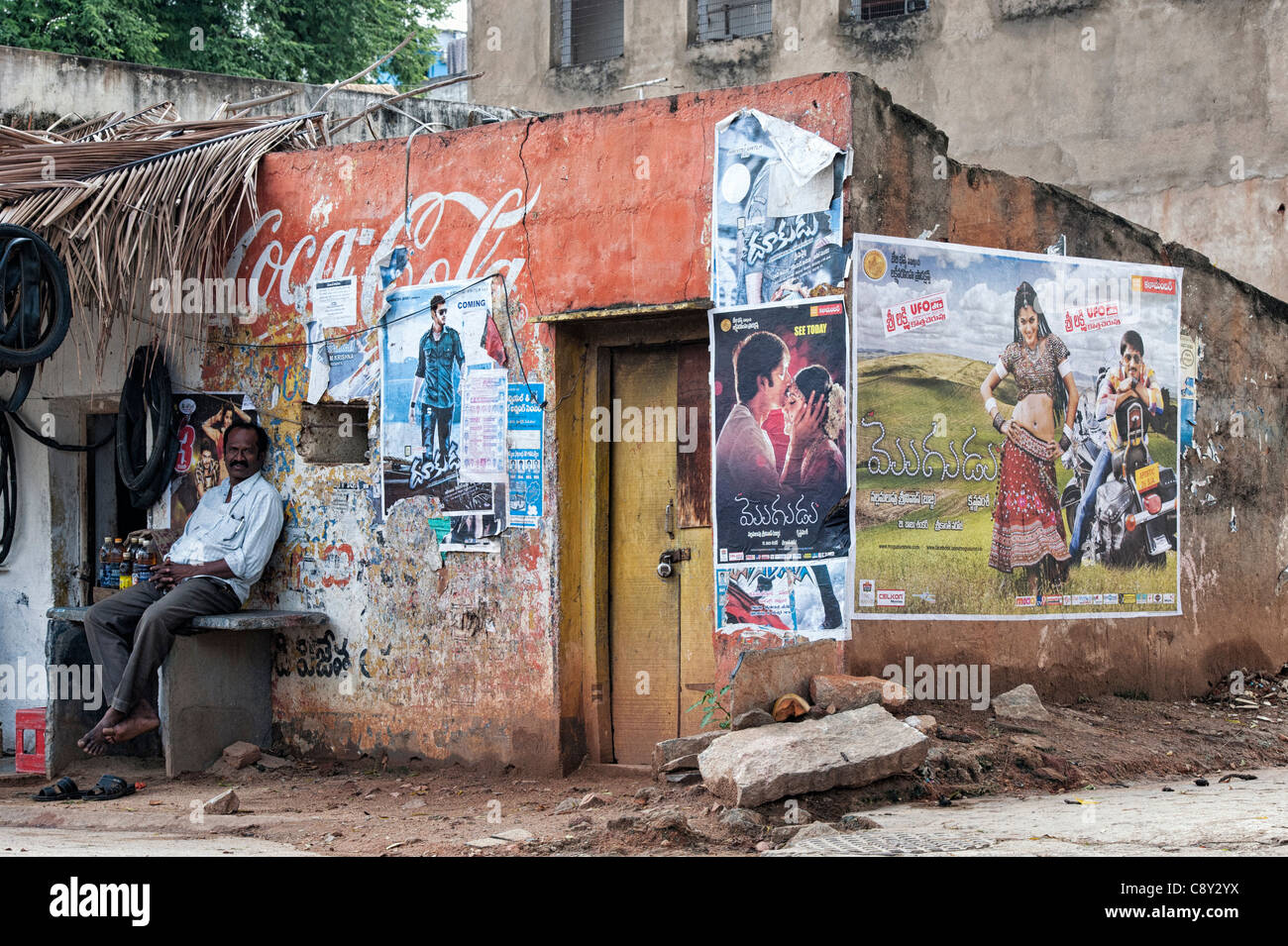 L'Inde homme assis à l'extérieur d'un mur en lambeaux d'une vieille maison couverte de l'Inde rurale indienne en affiches. Puttaparthi, Andhra Pradesh, Inde Banque D'Images