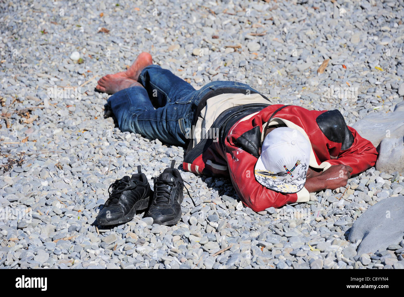 Un demandeur d'asile se trouvant sur une plage de galets avec ses chaussures à côté de lui Banque D'Images