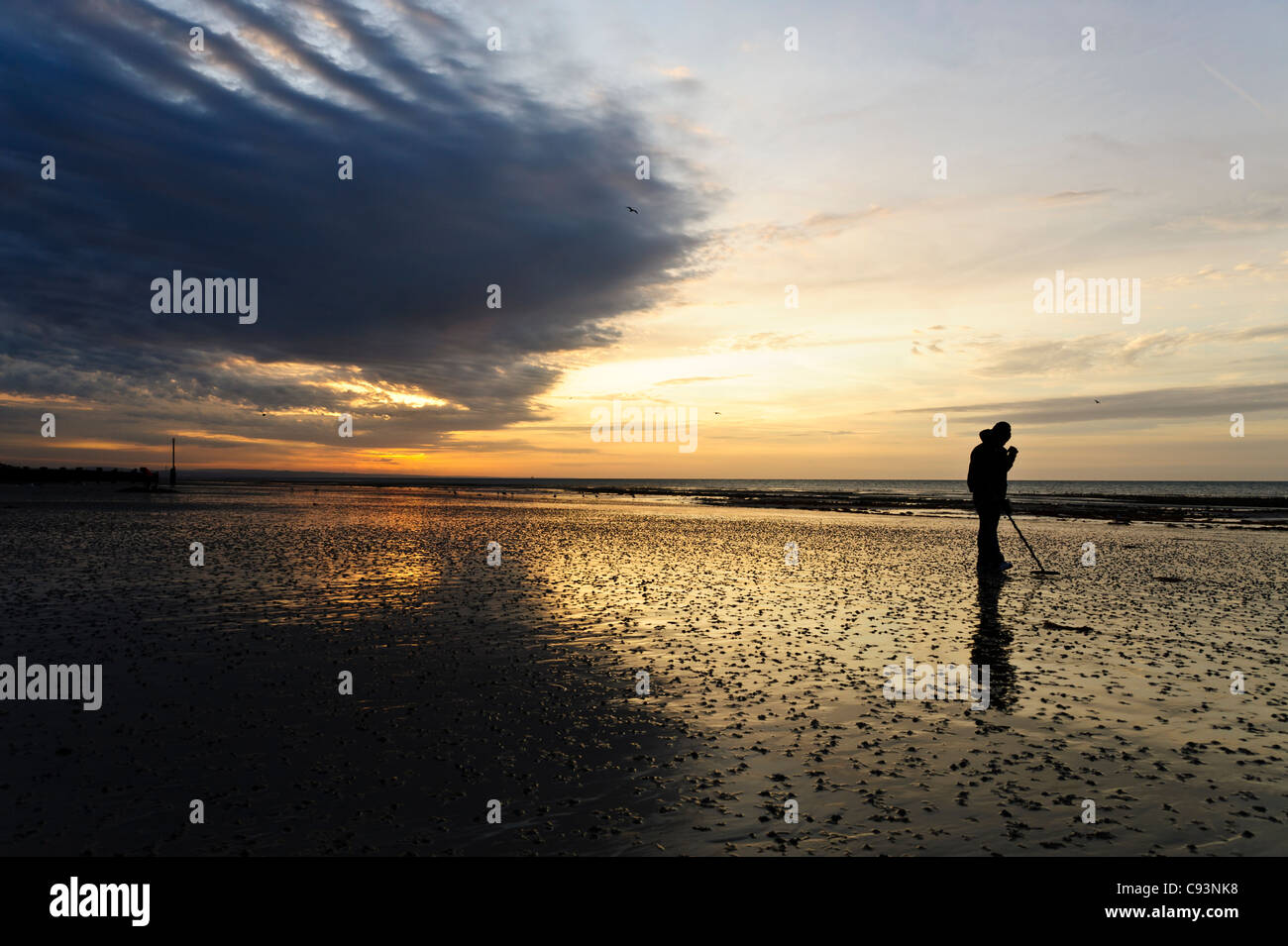 Un homme en peignant sur la plage de Worthing Beach en utilisant un détecteur de métal au lever du soleil. Banque D'Images