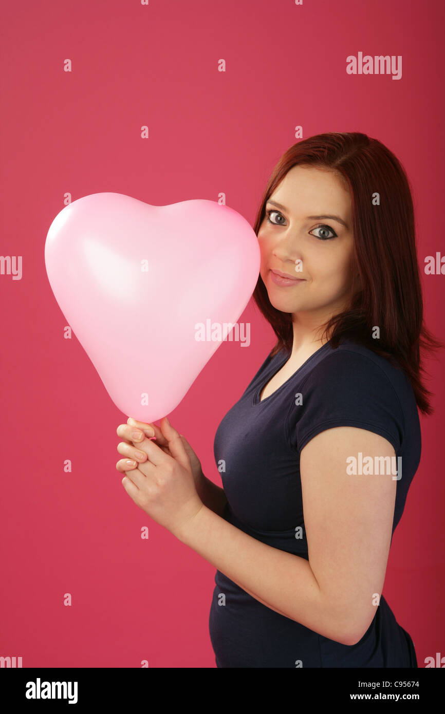 Jeune femme tenant un ballon en forme de coeur rose à côté de son visage. Banque D'Images