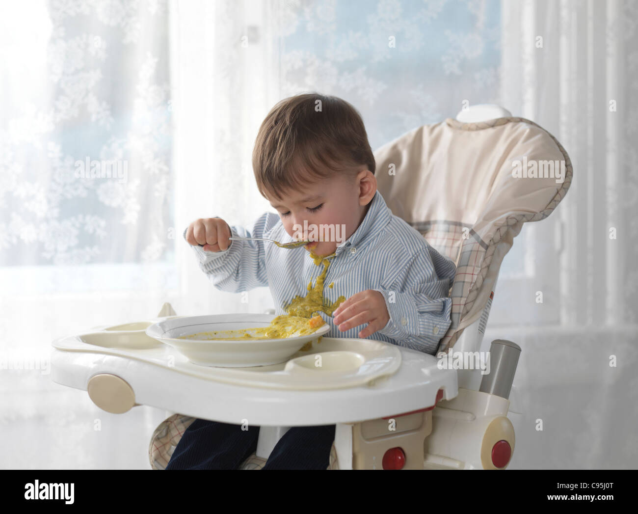 Un an et demi ans garçon assis sur une chaise haute et de manger la soupe avec une cuillère, déversant sur son T-shirt Banque D'Images