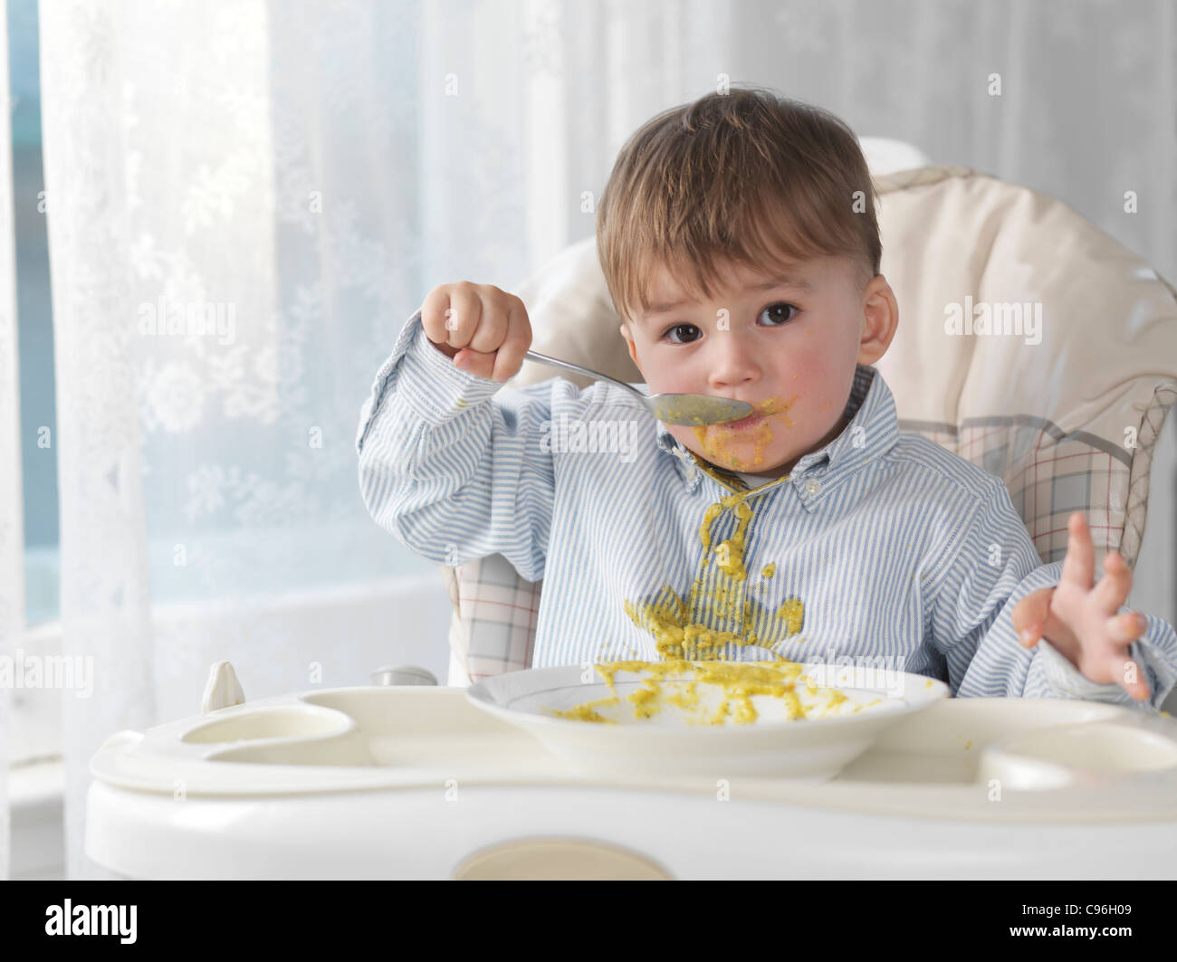 Mignon bébé garçon assis sur une chaise haute et de manger la soupe avec une cuillère, déversant sur son T-shirt Banque D'Images