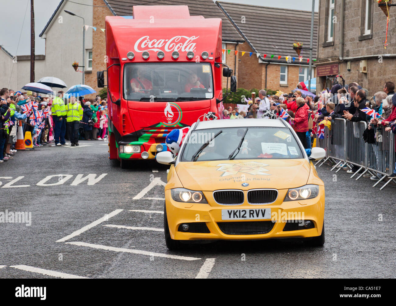 Les véhicules de soutien de six minutes d'avance sur la flamme olympique lors de son passage dans le village de Barrmill dans Ayrshire du Nord le 8 juin 2012. Thomas de flambeau, Tracey, 59 (près du centre) attend que la flamme d'être transmis à sa torche. Banque D'Images