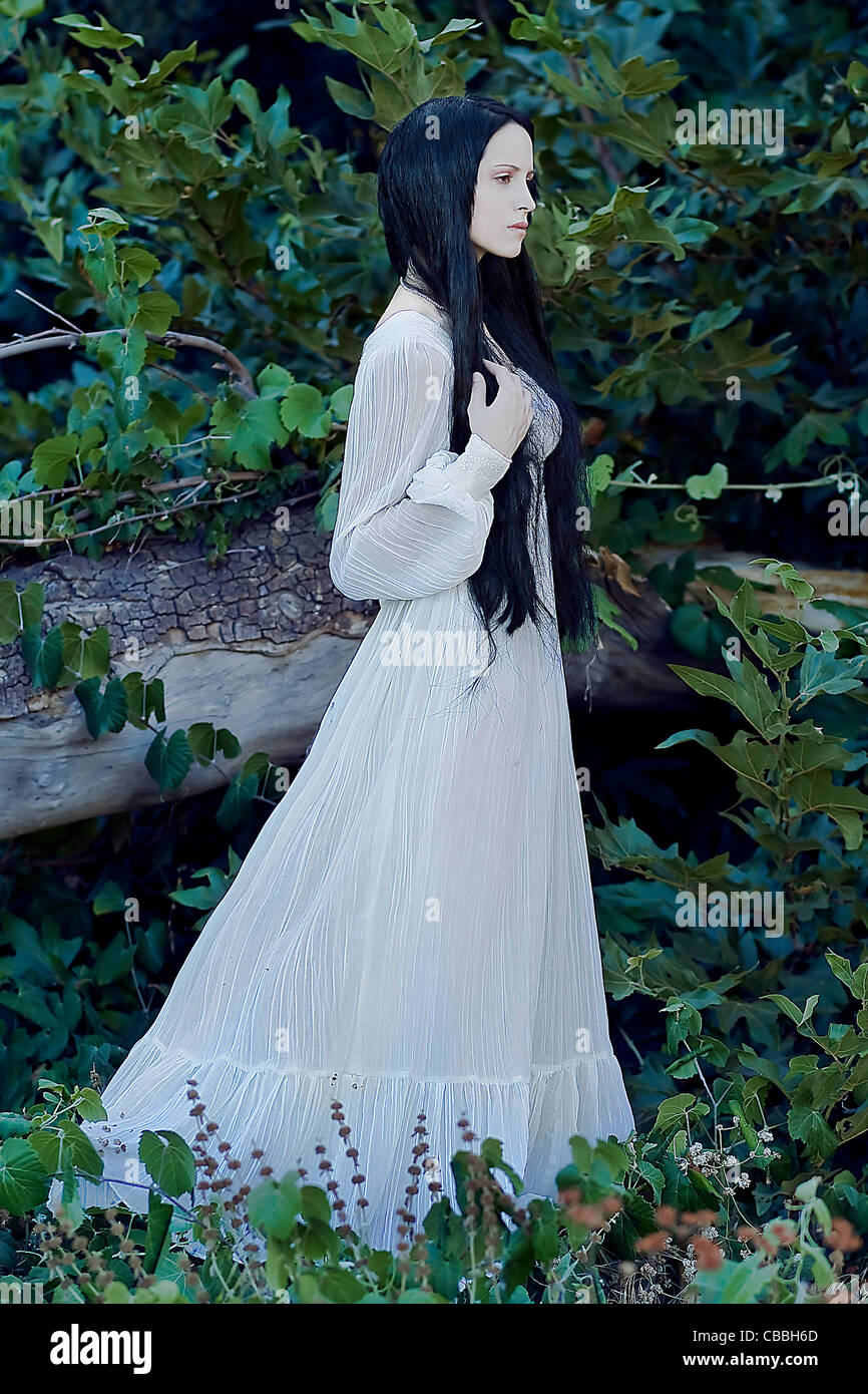 Une jeune femme aux longs cheveux noirs portant une longue robe blanche à l'extérieur à côté d'un arbre tombé sentir victorienne Banque D'Images