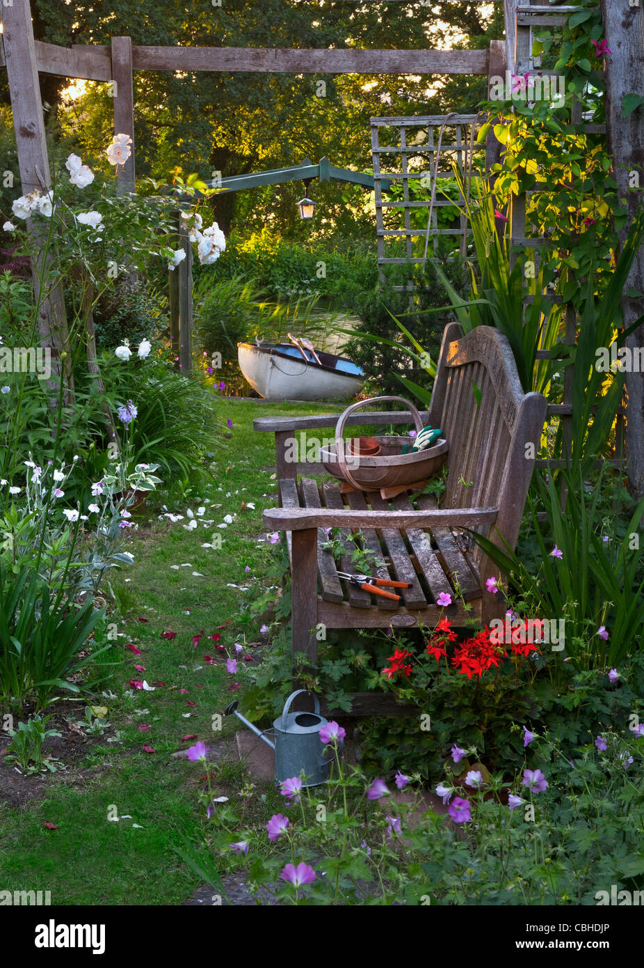 banc de jardin et roses sous la périgola dans le jardin floral avec des outils de jardinage de trug traditionnels bateau à ramer et rivière derrière au coucher du soleil Banque D'Images