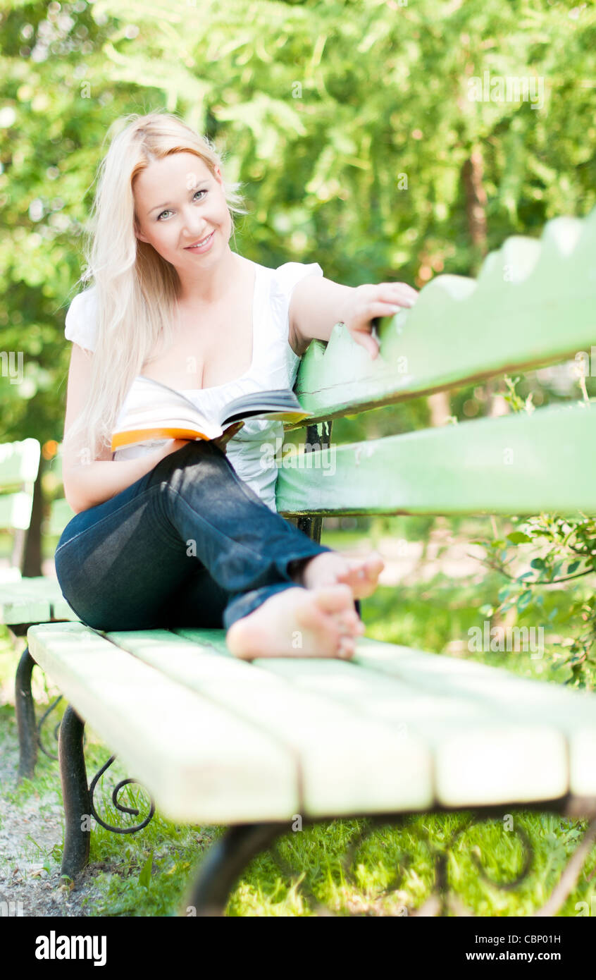 Jeune femme assise sur un banc dans le parc, reading book, souriant et à la recherche dans l'appareil photo Banque D'Images