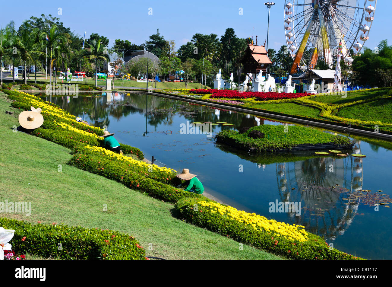 Jardiniers avec d'énormes chapeaux faisant l'entretien paysager à côté d'étang à l'expo à Chiang Mai en Thaïlande avec la grande roue en arrière-plan Banque D'Images