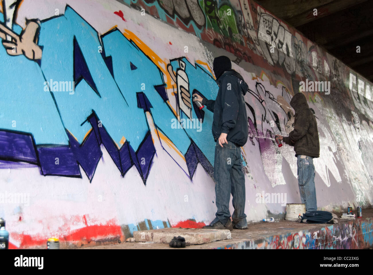 Étiquetage - Deux jeunes hommes cagoulés graffiti peinture par pulvérisation sous le pont, à l'ouest de Londres, Royaume-Uni Banque D'Images