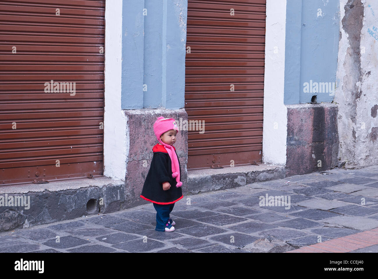 Un enfant se trouve sur un trottoir en face d'un magasin fermé en hiver, vêtements de laine rose, bouchon fantaisie manteau noir avec garniture rouge. Banque D'Images