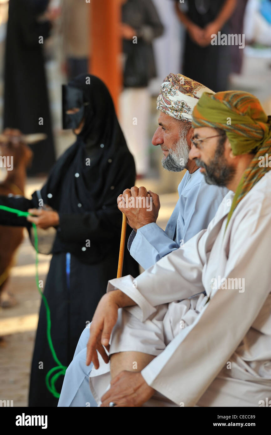 Deux omanis regarder la foule tandis qu'une femme vêtue de noir avec un masque de se tenir en arrière plan à Nizwa Oman ; marché de la chèvre. Banque D'Images