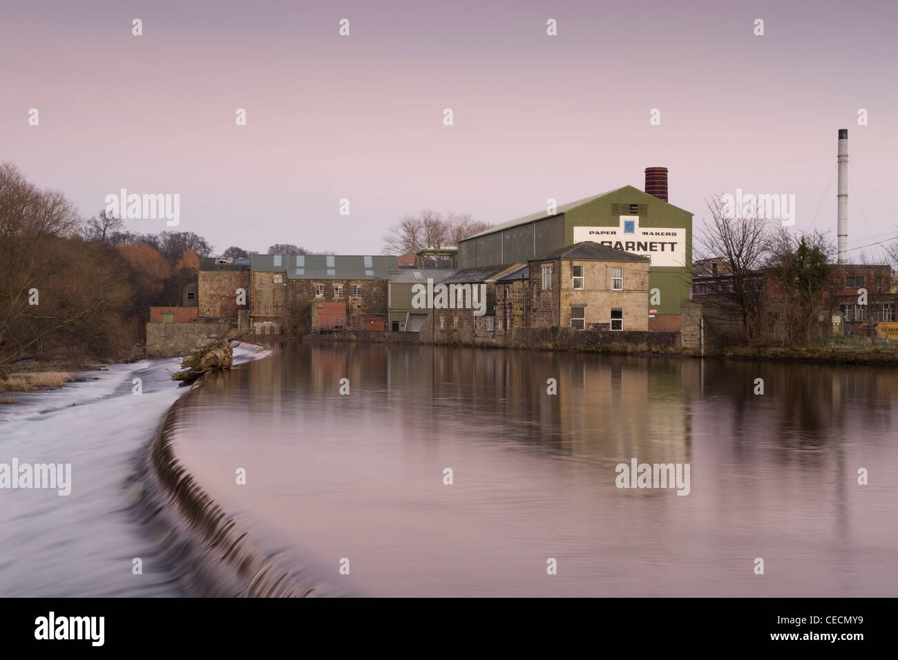 L'eau qui coule en cascade de River Wharfe sur weir sous ciel coucher de soleil rose, grenat historique au-delà du moulin à papier - Otley, West Yorkshire, Angleterre, Royaume-Uni. Banque D'Images