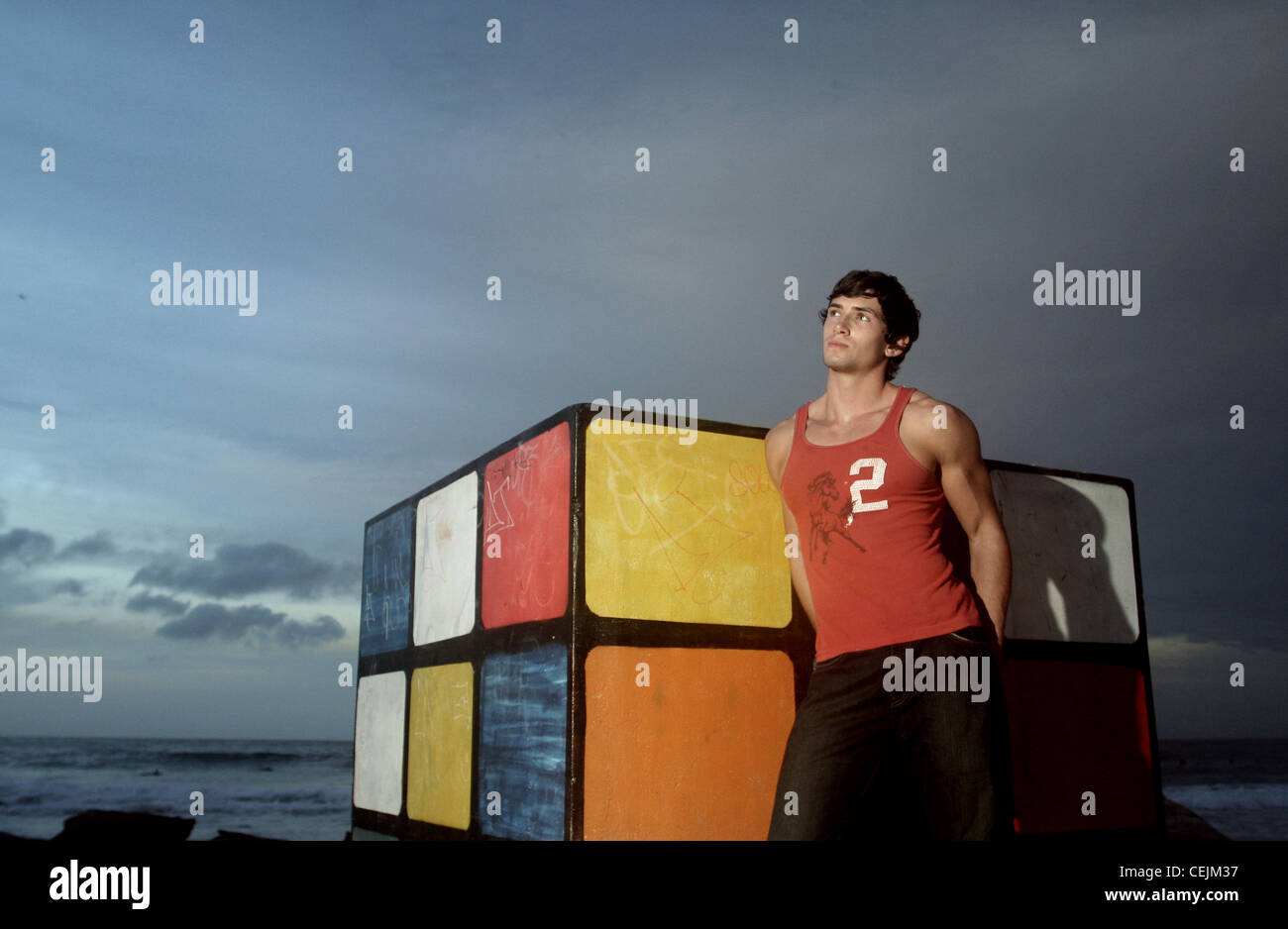 Brune, les cheveux des hommes portant des jeans noir et veste rouge, debout sur la plage, s'appuyant contre le Rubiks Cube, souriant, Banque D'Images