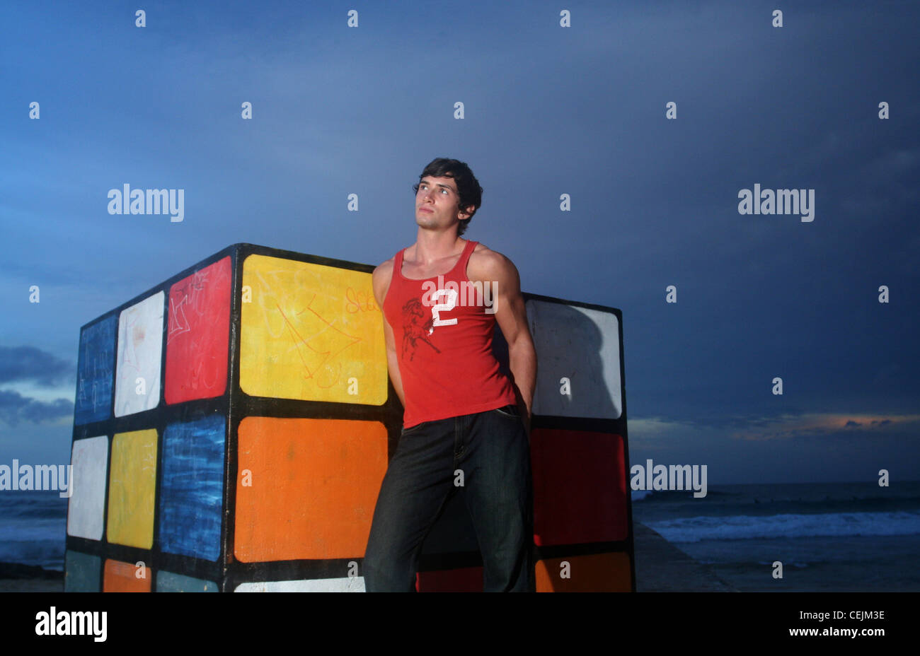 Brune, les cheveux des hommes portant des jeans noir et veste rouge, debout sur la plage, s'appuyant contre le Rubiks Cube, souriant, Banque D'Images