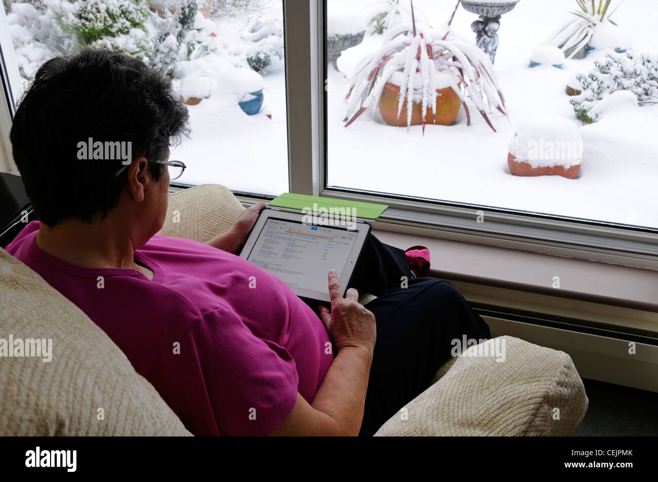 Femme sénior mature titulaire d'un Apple iPad tablette numérique sélection d'applications par fenêtre assis dans un fauteuil hiver neige dehors Angleterre Royaume-Uni Banque D'Images