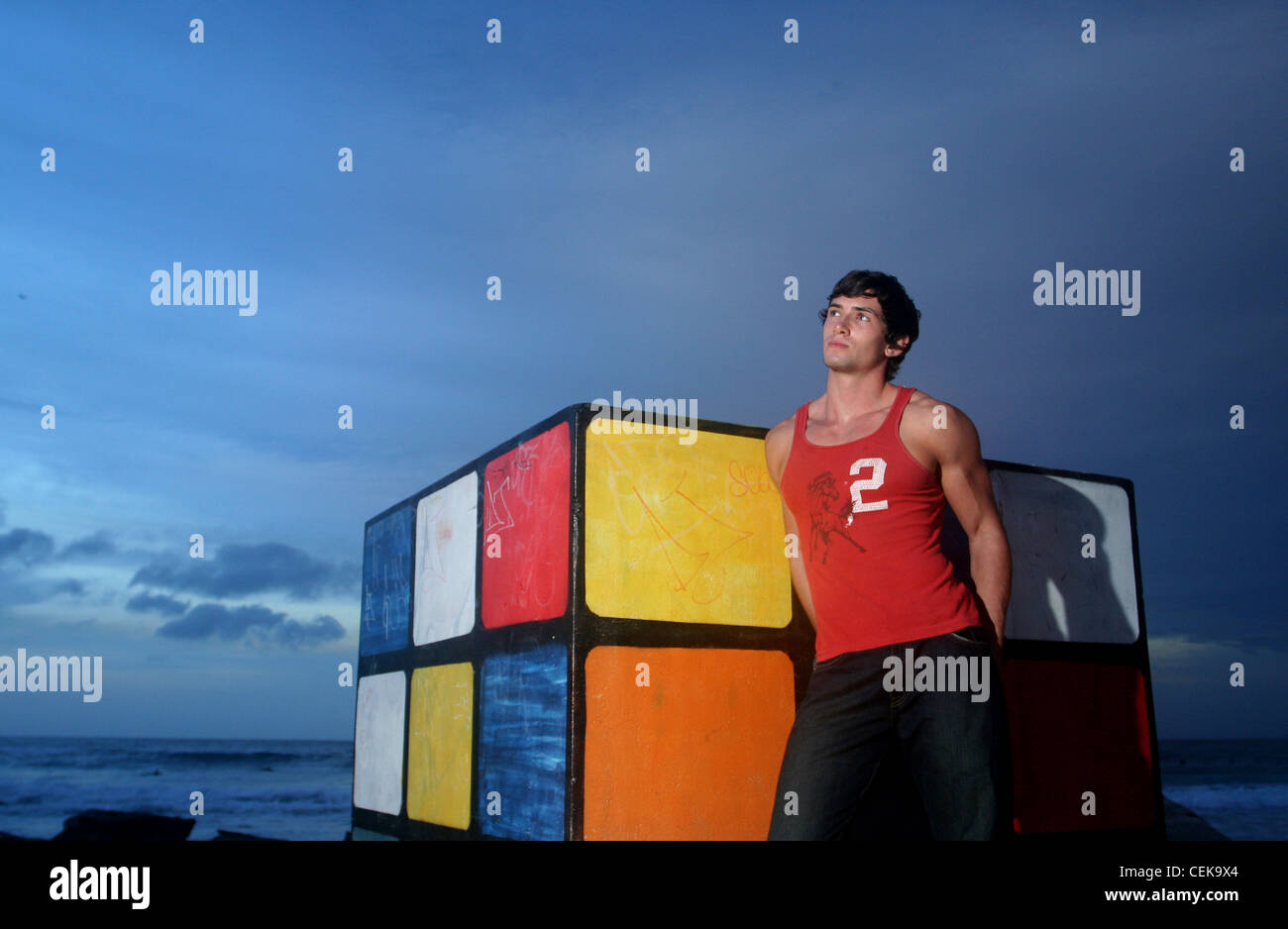 Brune, les cheveux des hommes portant des jeans noir et veste rouge, debout sur la plage, s'appuyant contre le Rubiks Cube, souriant, Banque D'Images