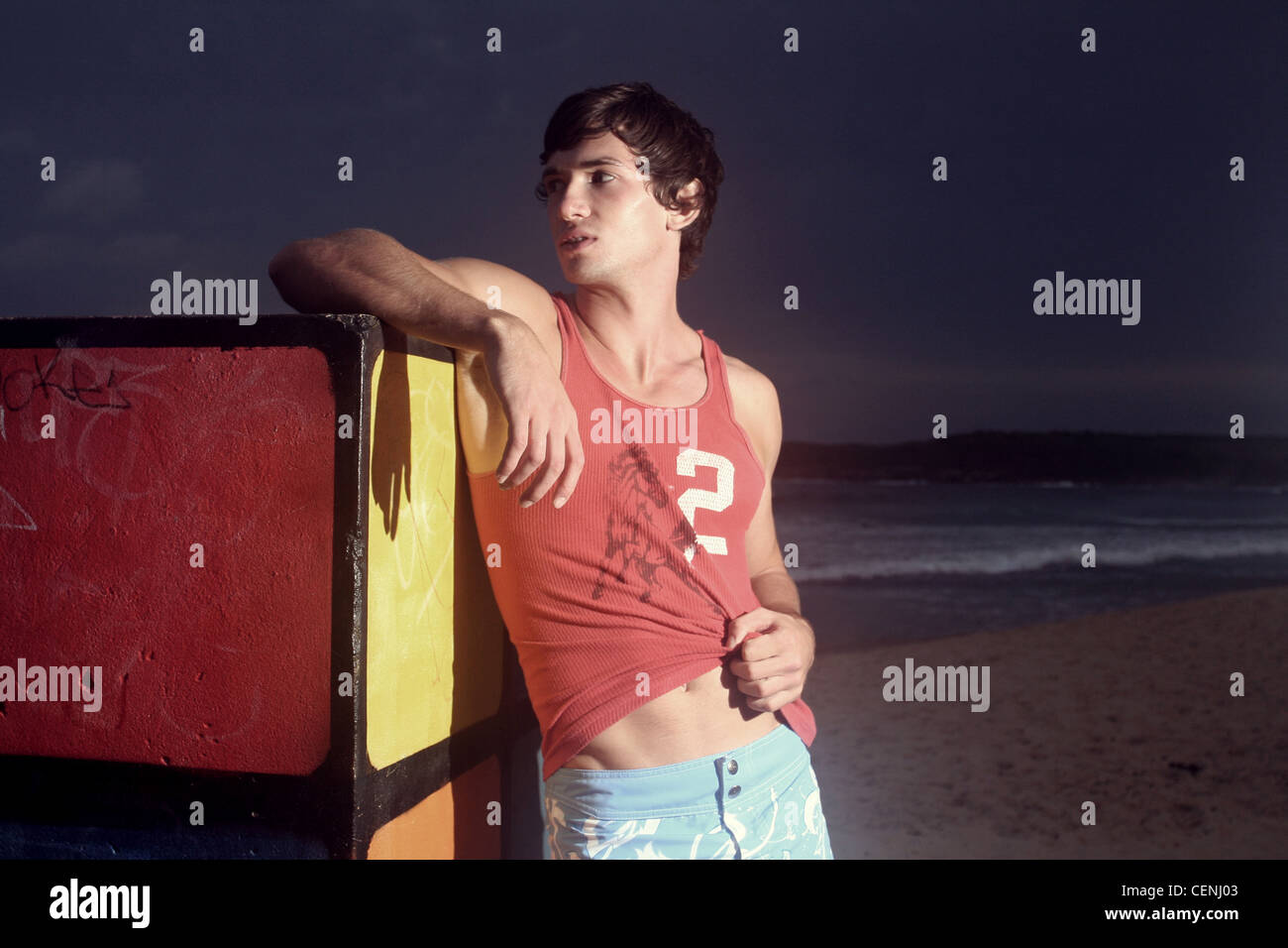 Brune, les cheveux des hommes portant des shorts bleu et veste rouge, debout sur la plage, s'appuyant contre le Rubiks Cube, souriant, Banque D'Images