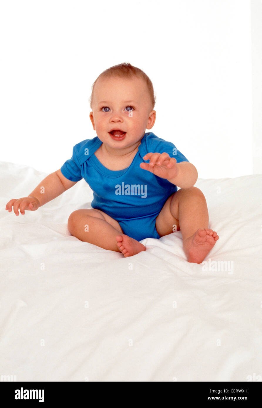 Bébé mâle aux yeux bleus assis avec ses bras tendus sur une couette blanche sur un lit Banque D'Images