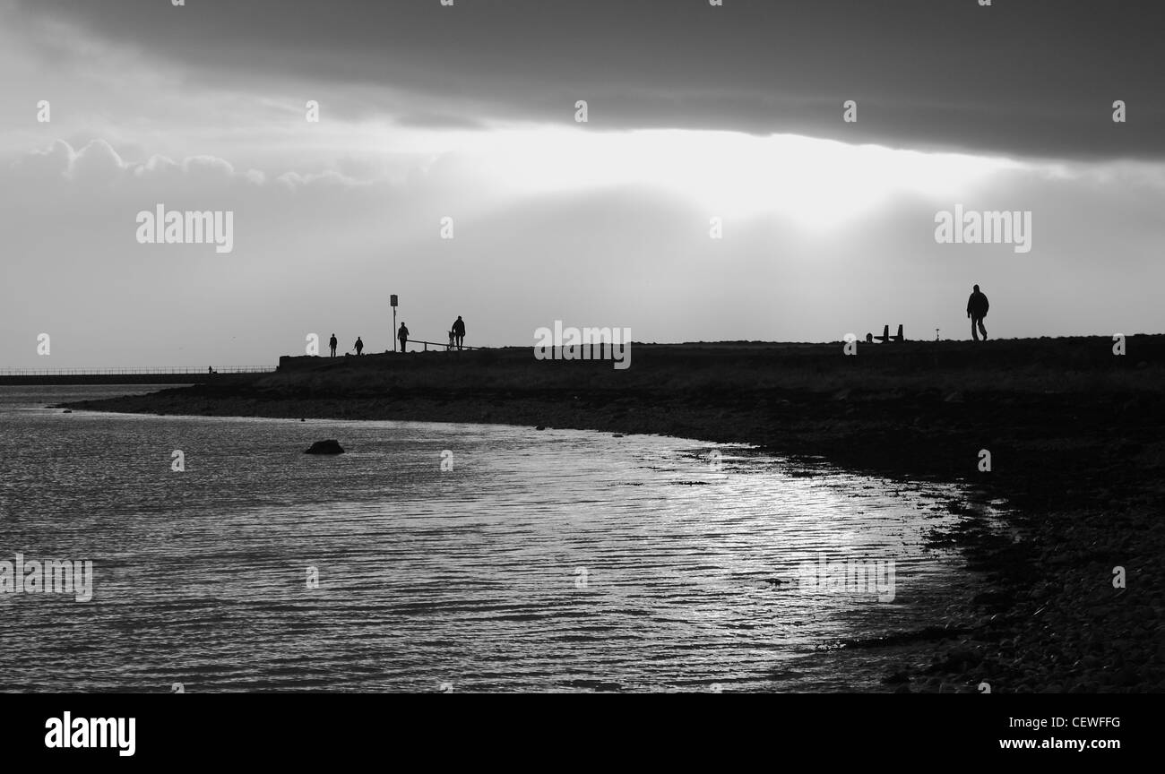 Un tir de personnes à pied le long du littoral de la baie de Galway vers le soleil percer les nuages créant des silhouettes. Banque D'Images