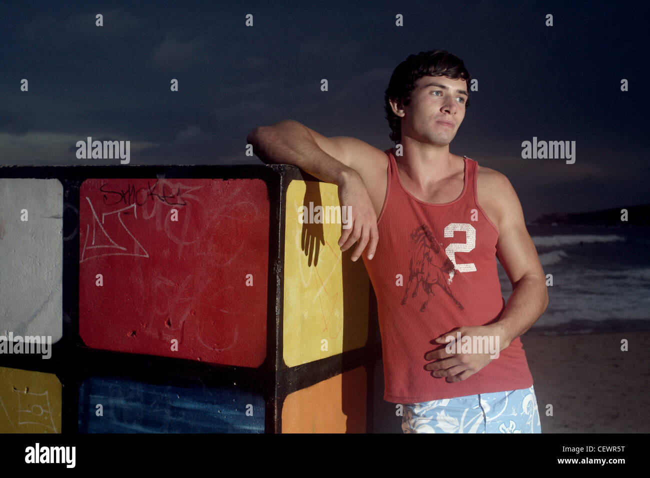 Brune, les cheveux des hommes portant des shorts bleu et veste rouge, debout sur la plage, s'appuyant contre le Rubiks Cube, souriant, Banque D'Images