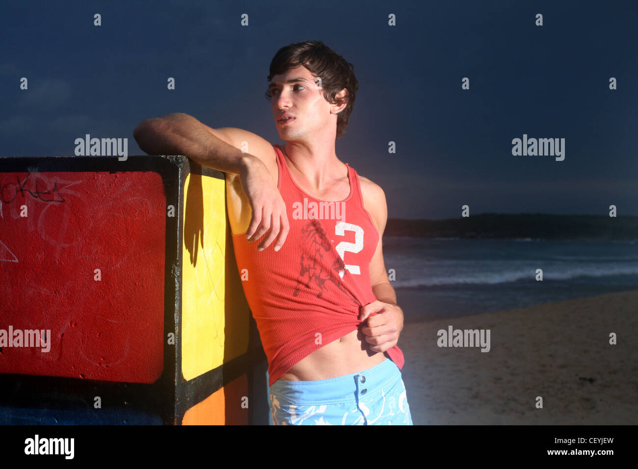 Brune, les cheveux des hommes portant des shorts bleu et veste rouge, debout sur la plage, s'appuyant contre le Rubiks Cube, souriant, Banque D'Images