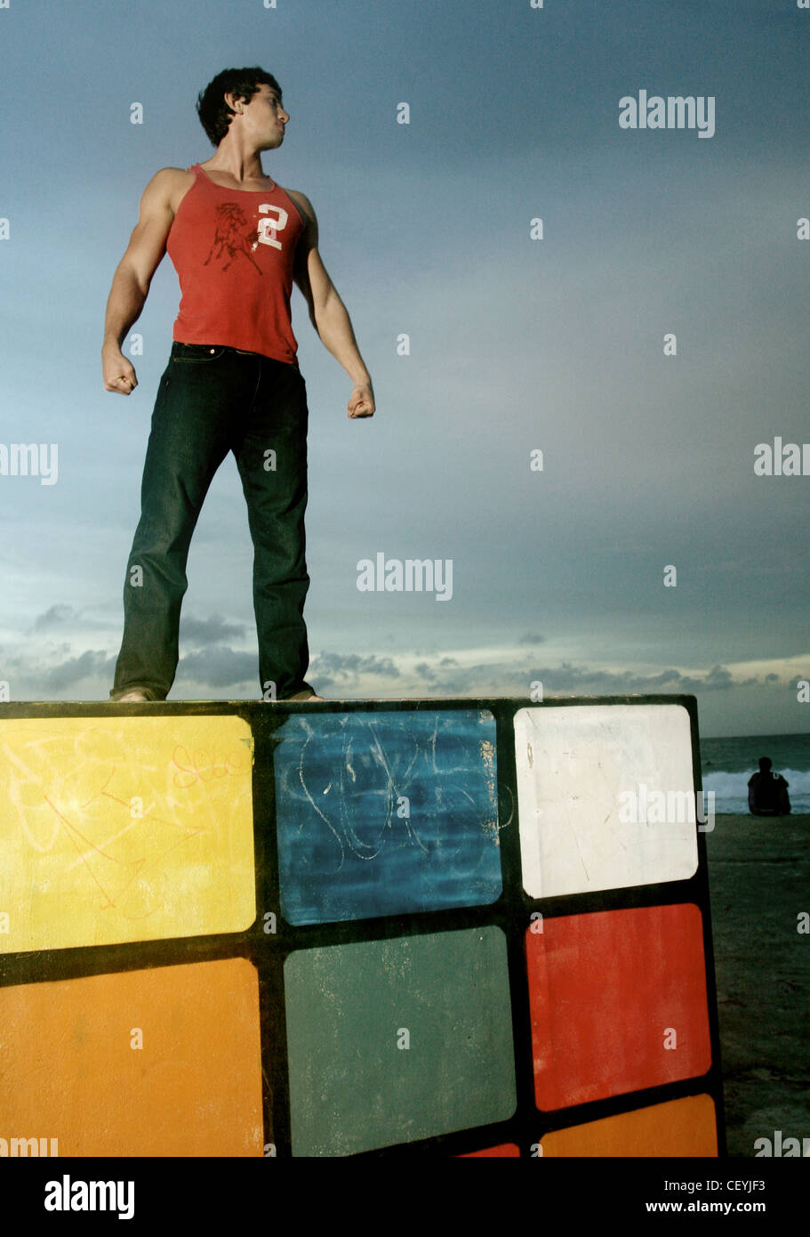 Brune, les cheveux des hommes portant des jeans noir et veste rouge, debout sur la plage sur le dessus de la Rubiks Cube, souriant, à la recherche de Banque D'Images