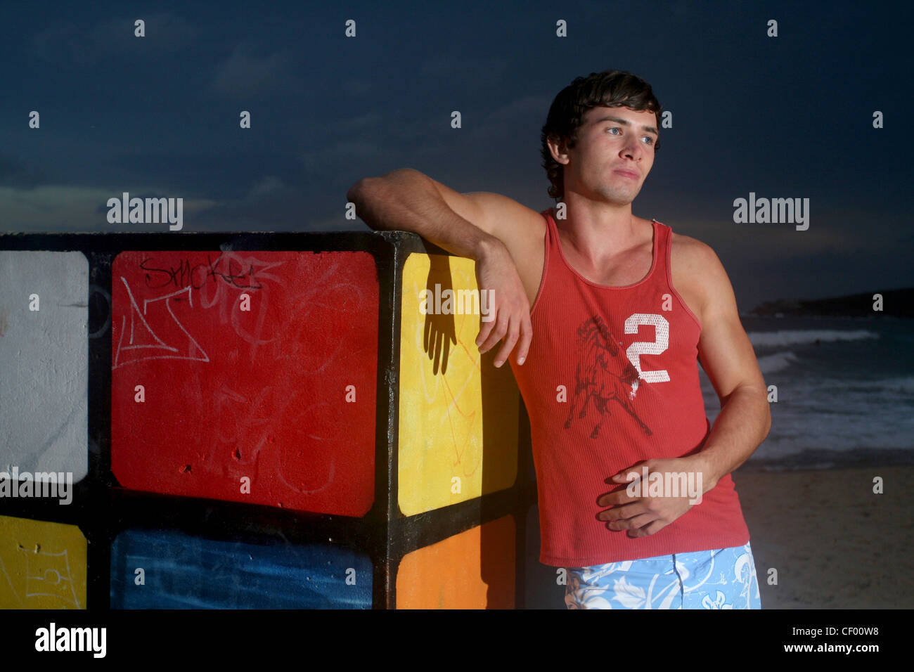 Brune, les cheveux des hommes portant des shorts bleu et veste rouge, debout sur la plage, s'appuyant contre le Rubiks Cube, souriant, Banque D'Images