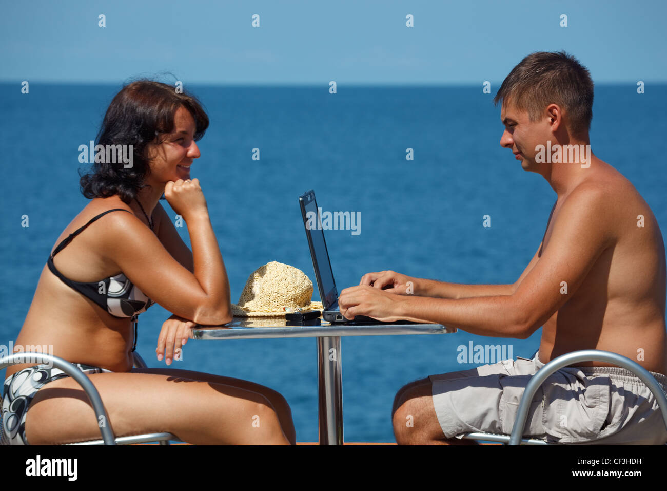 L'homme et la femme assis à table sous open sky sur fond de mer. L'homme travaille pour ordinateur portable. Banque D'Images