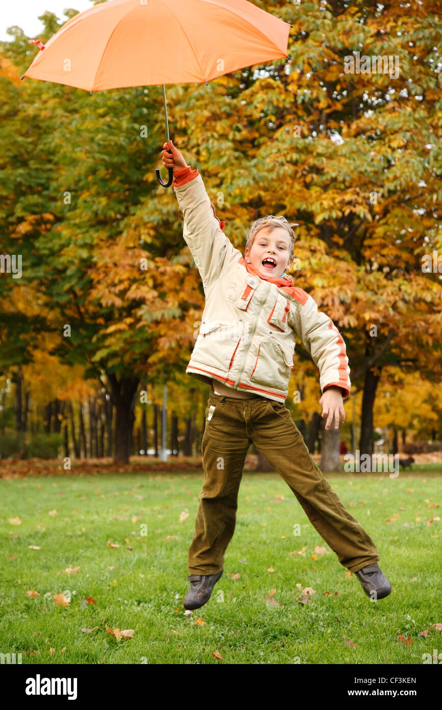 Happy boy in autumn park. A sauté avec parapluie orange sur la tête. Banque D'Images