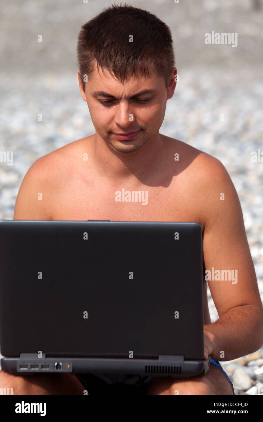 Jeune homme travaille sur son ordinateur portable sur littoral. Homme avec portable se trouve sur une plage de galets. Banque D'Images