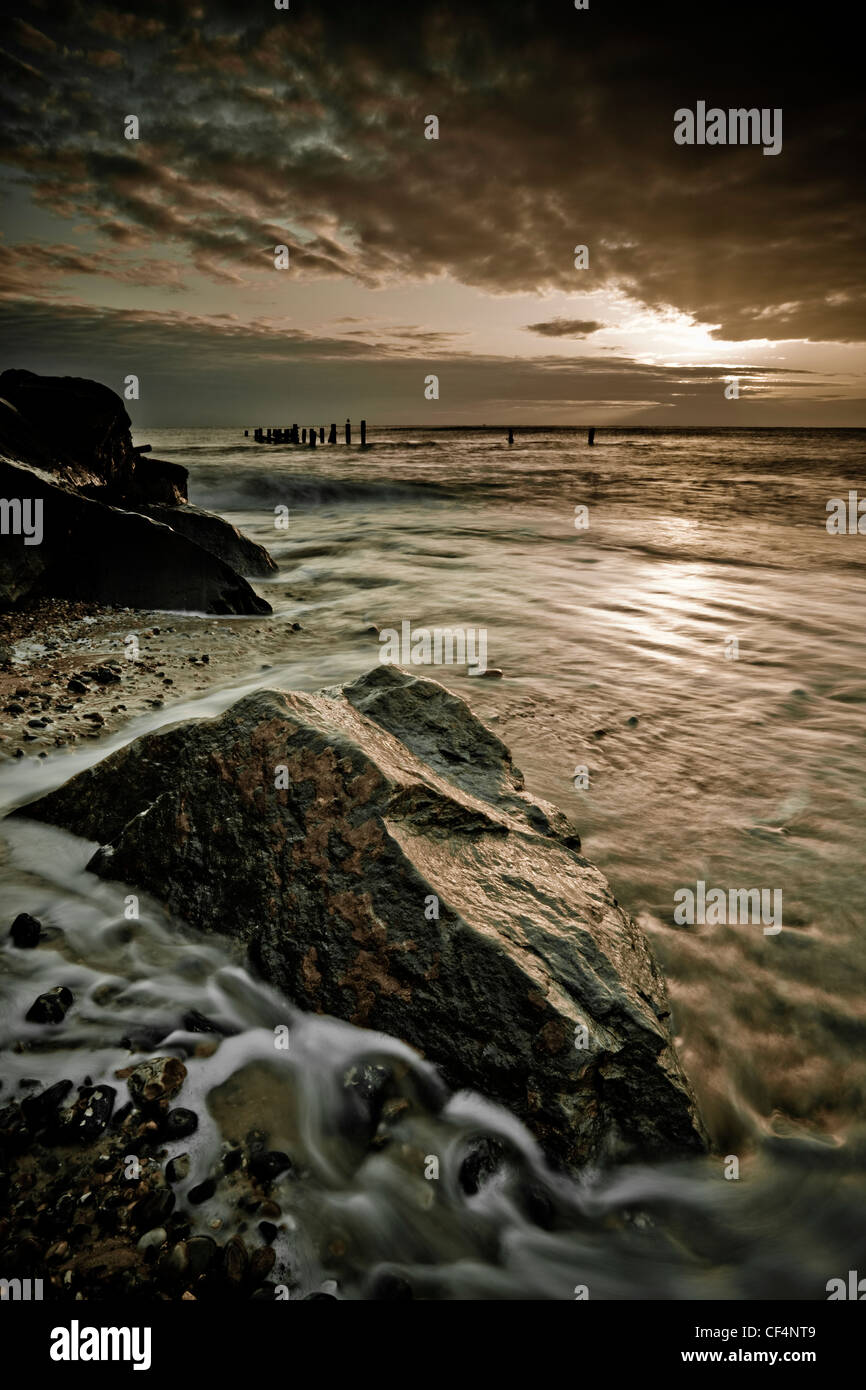 Briser le soleil nuages pendant le lever du soleil sur les gros rochers sur la plage à Happisburgh sur la côte nord-est de Norfolk. Banque D'Images