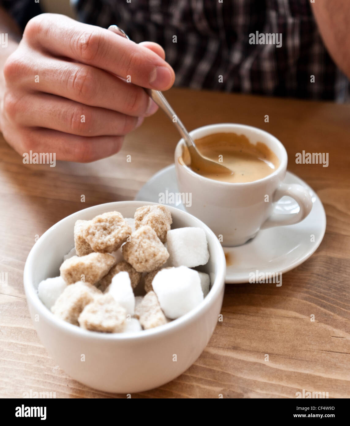 Détail de la main de l'homme tenant une cuillère en remuant une tasse de café expresso, avec un bol de sucre et des morceaux de sucre en premier plan. Banque D'Images