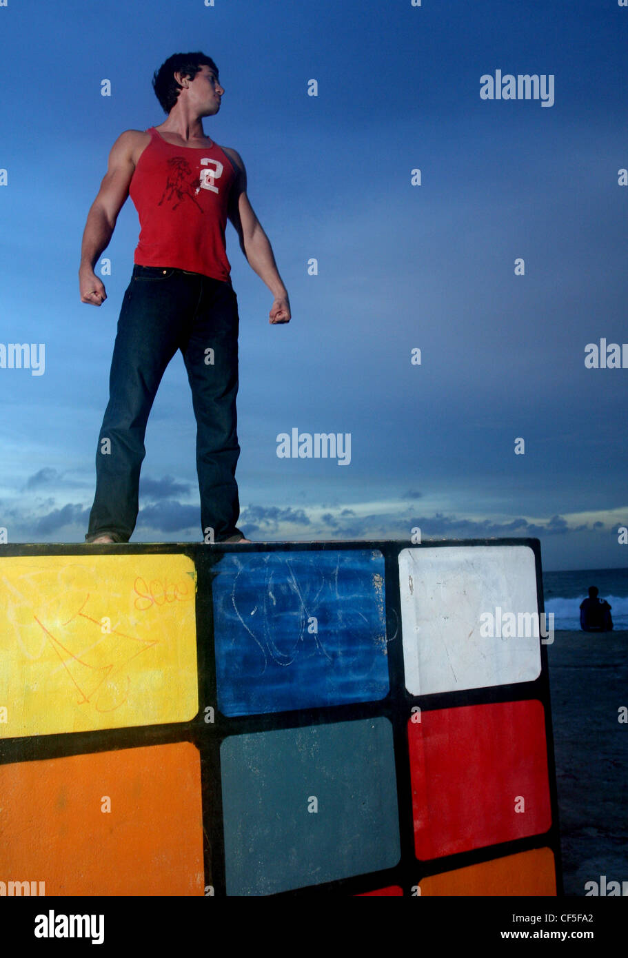 Brune, les cheveux des hommes portant des jeans noir et veste rouge, debout sur la plage sur le dessus de la Rubiks Cube, souriant, à la recherche de Banque D'Images
