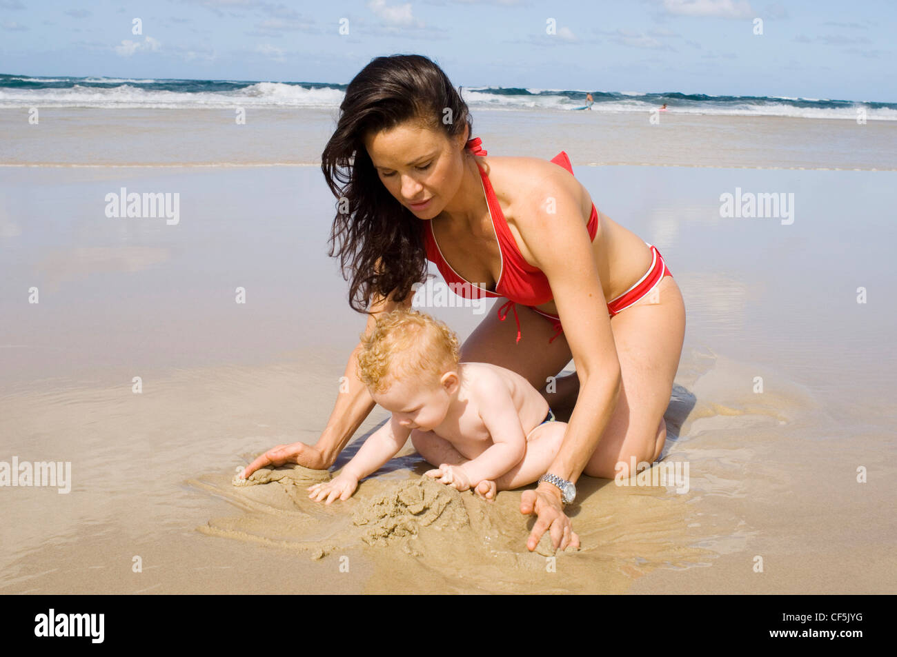Femme brune long cheveux, wearing red bikini, à genoux sur une plage de  sable, bébé de sexe masculin cheveux blonds, portant un pantalon bleu  piscine Photo Stock - Alamy