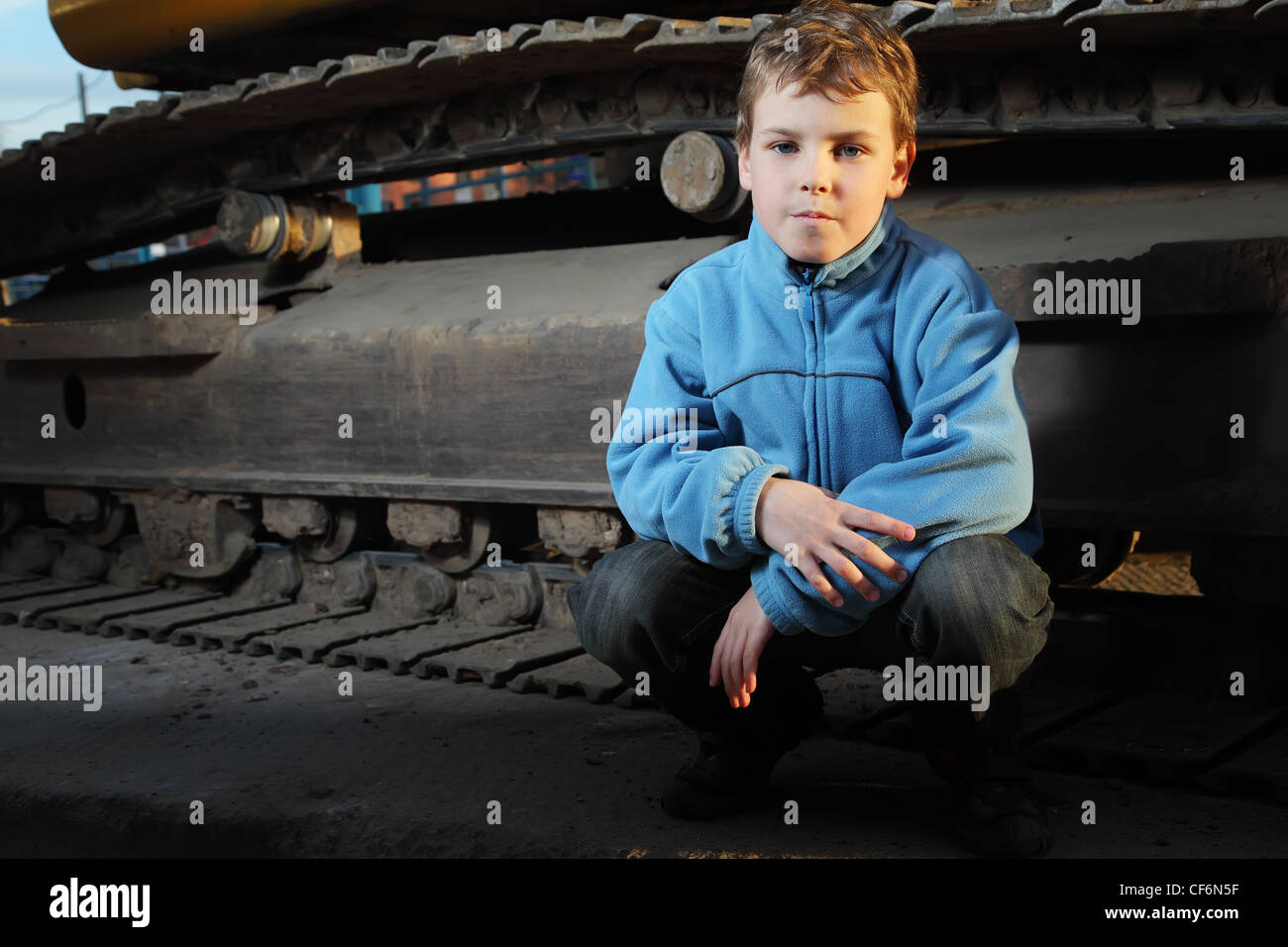 Petit garçon en veste bleue assis près de tracteur à chenilles dans le noir Banque D'Images