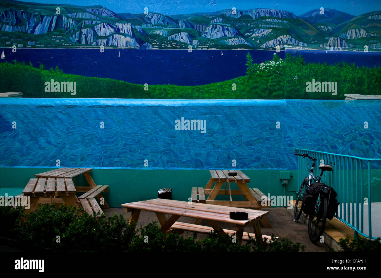 Café terrasse avec tables et banc un vélo contre un mur avec un mur peint avec un paysage de mer et montagne Banque D'Images