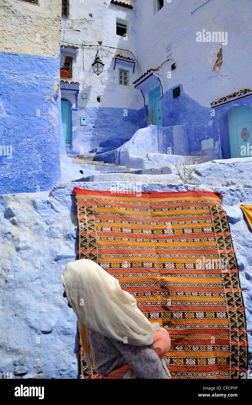 Vieille Femme en face d'un tapis et des murs bleus dans une ruelle étroite à Chefchaouen, Maroc, montagnes Riff, Afrique Banque D'Images